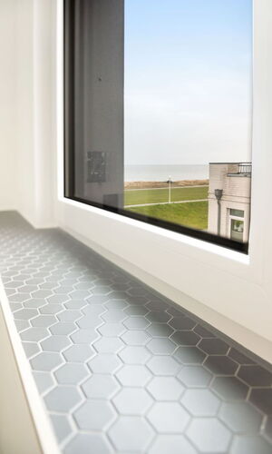 Fensterbank mit grauen sechseckigen Fliesen, Blick auf Rasenfläche und Gebäude mit Meer im Hintergrund